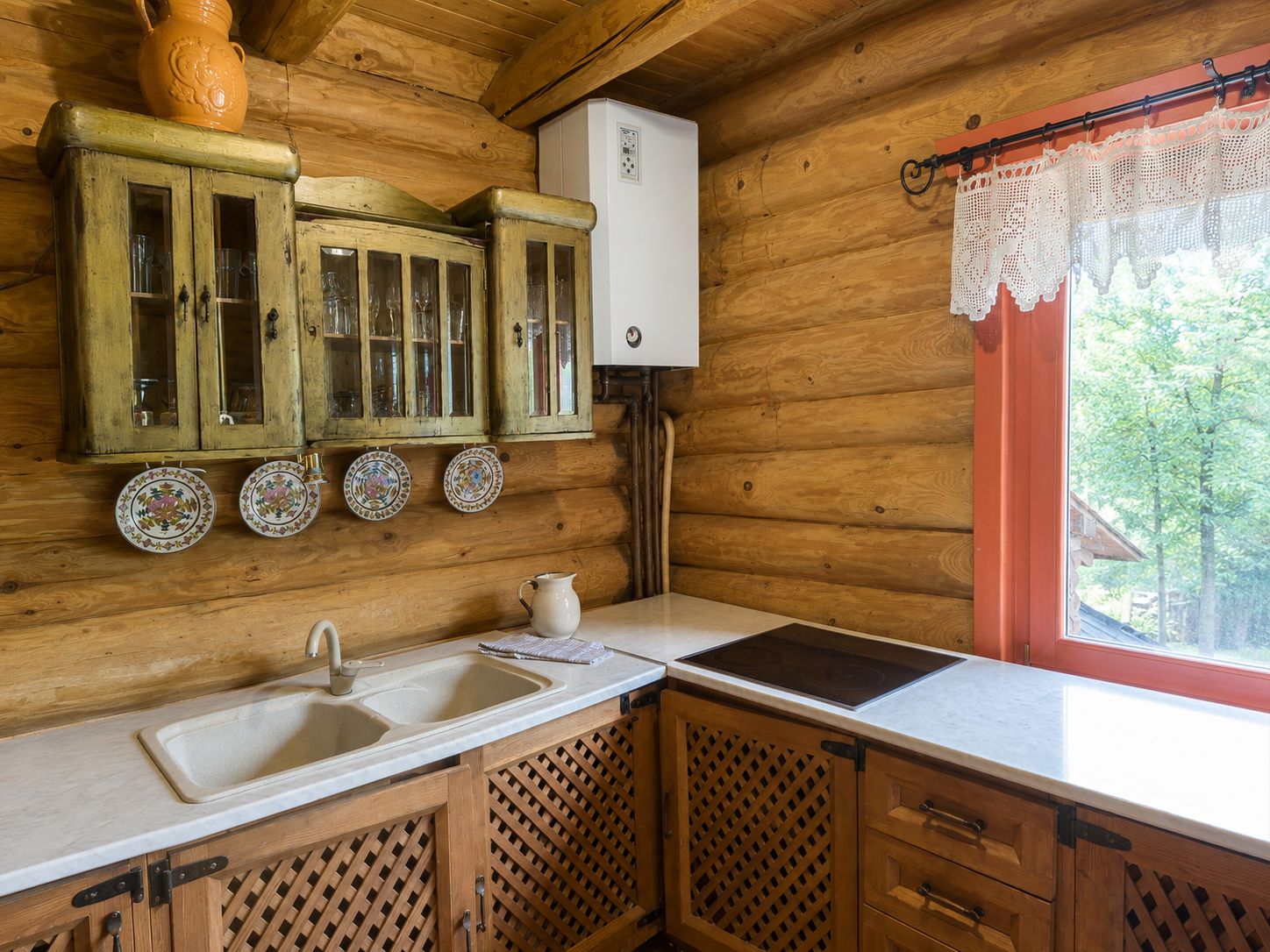 A wooden cabin interior with a kitchen area, including a sink, cabinets, and a window with lace curtains.