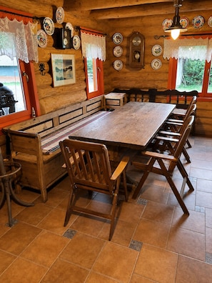 A wooden dining area with a long table and benches, a clock on the wall, and a window with lace curtains. - Traditional Wooden Cottage with private garden situated in Campu Cetatii (Câmpu Cetăţii)