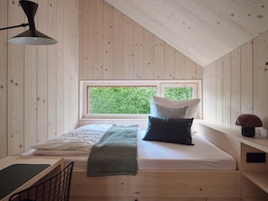 A bedroom with a sloped ceiling, a window with a view of greenery, a bed with pillows, a bedside table, and a lamp. - Seeberg Homes - House 2 (Schondorf am Ammersee)