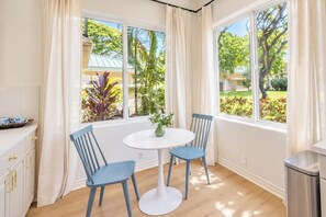 A room with a round table, two chairs, a potted plant, and a view of a garden through large windows. - The Azure at Mauna Kea: Relaxed Luxury + Ocean Views + Resort Option (Kamuela)