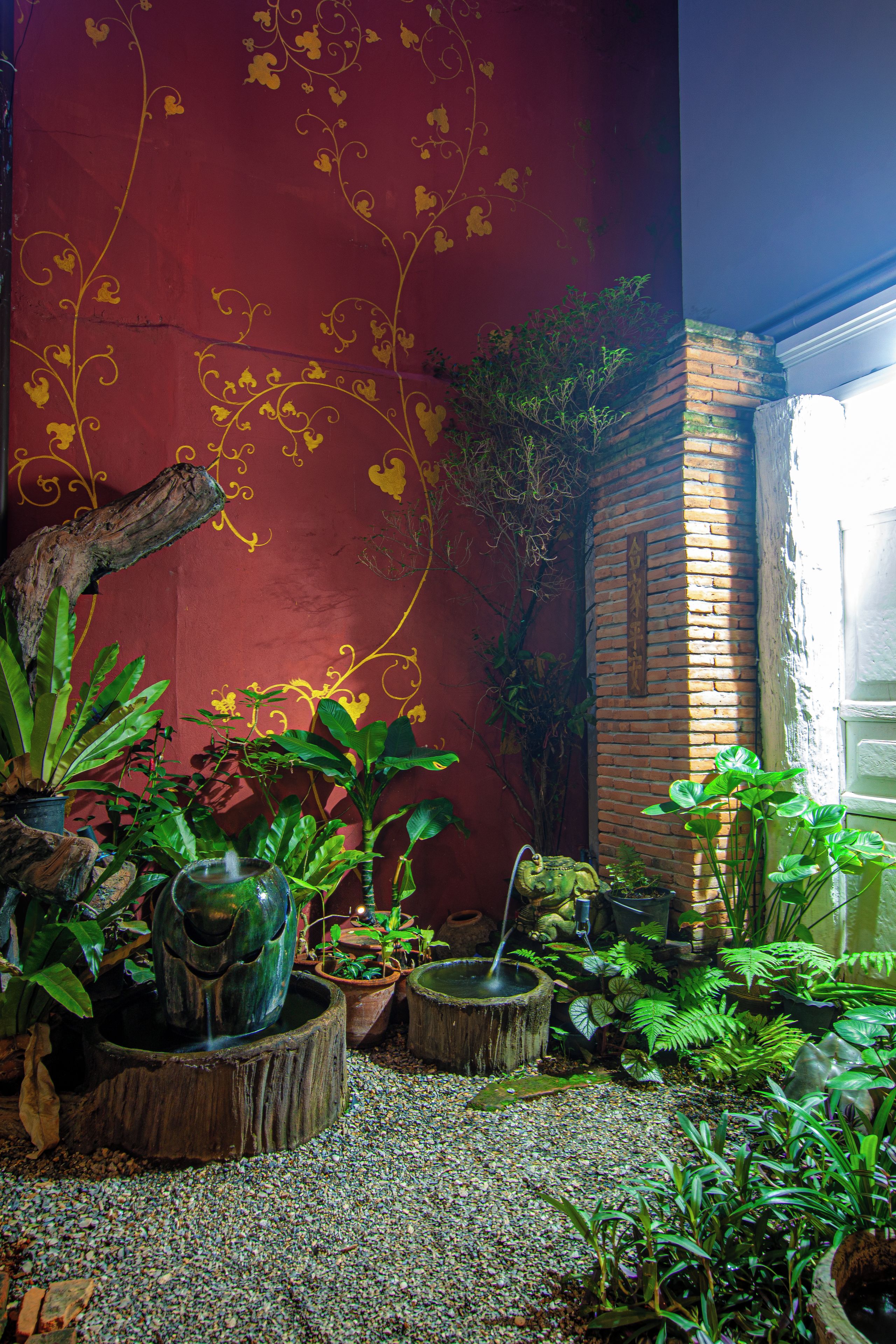 Une pièce avec un mur rouge, un mur en briques et une fenêtre. On y trouve différentes plantes en pot et une sculpture en bois.