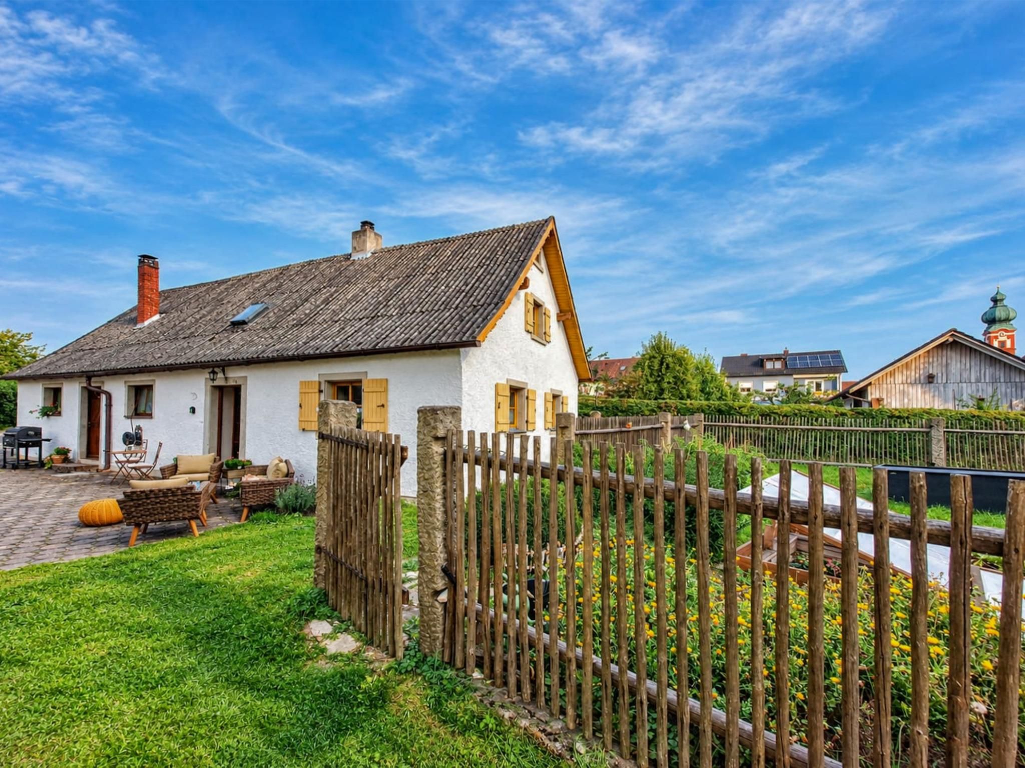A white house with a sloped roof, a wooden fence, and a garden with yellow flowers.