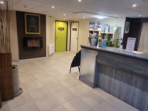 Een receptie met een receptiebalie, een ingelijst portret aan de muur en deuren met de aanduiding 'Chambres' en 'Toiletten'.