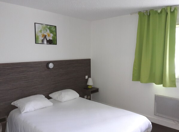Een hotelkamer met een bed, nachtkastje, lamp, wandklok en een raam met groene gordijnen.