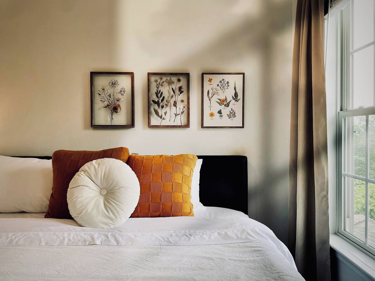 A bed with white bedding, a brown quilted pillow, and a white round pillow. There are three framed botanical prints on the wall above the bed.