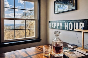 Une pièce avec une grande fenêtre, une carafe de whisky, des verres, des livres et un panneau indiquant « Happy Hour ».