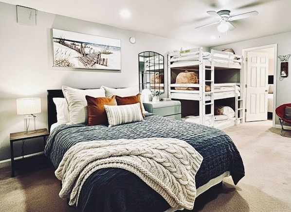 Une chambre à coucher comprenant un lit, des lits superposés, une table de chevet, une lampe, un ventilateur de plafond et une porte.