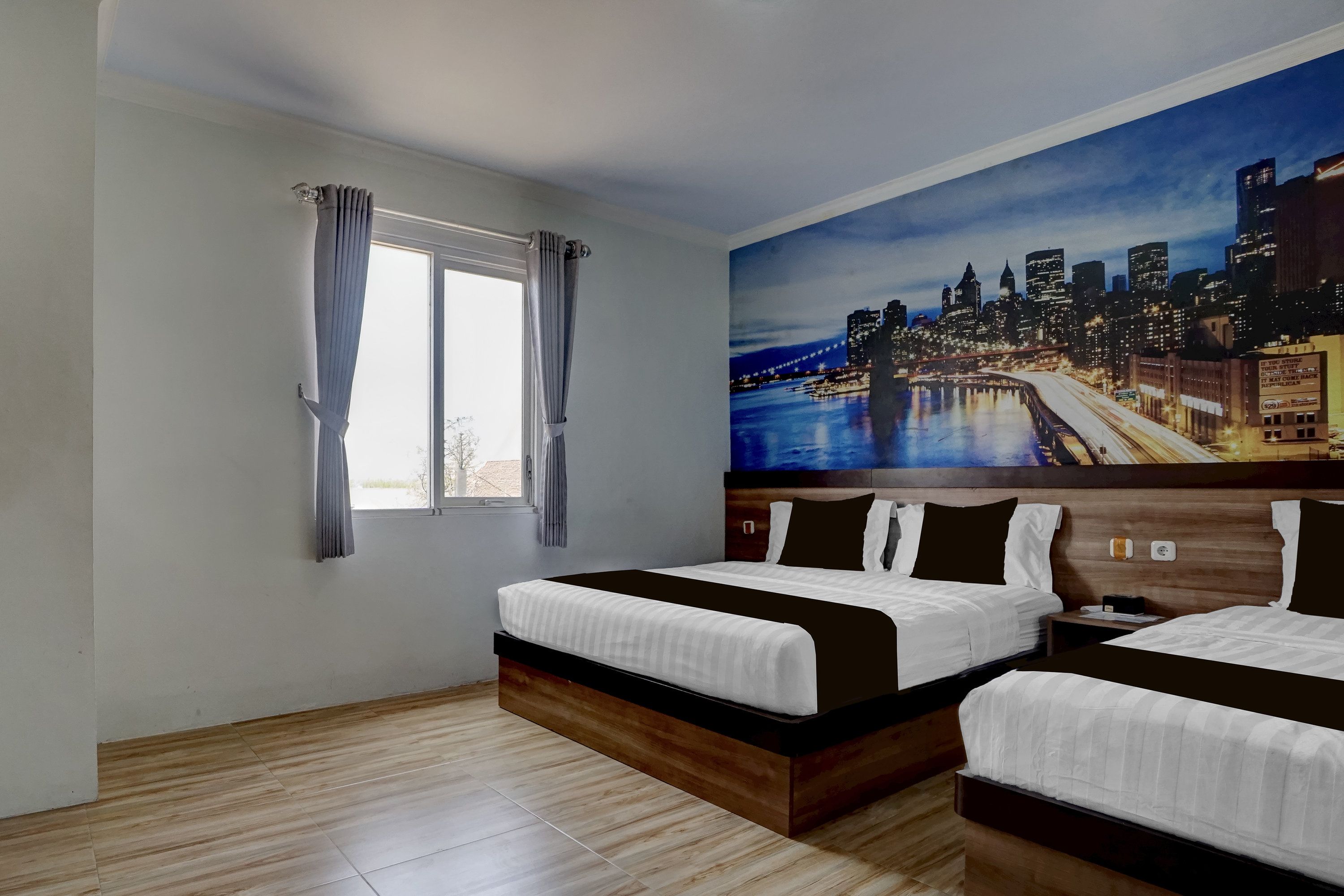 A hotel room with two beds, a large window with curtains, and a cityscape mural on the wall.