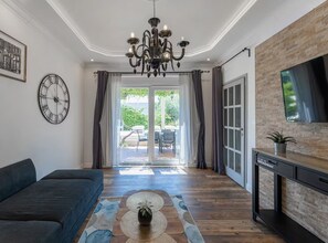 A living room with a dark sofa, a large clock, a chandelier, and a TV on a brick wall. - Deluxe Apartment Marghareta - Three Bedroom Apartment With Sea View and Terrace (Dubrovnik)