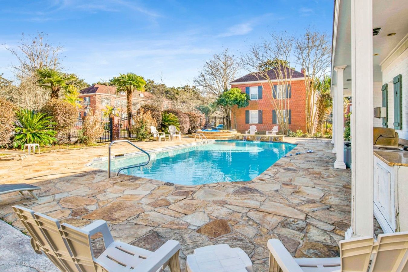 A swimming pool with stone tiles, surrounded by chairs and a building with a red brick facade.