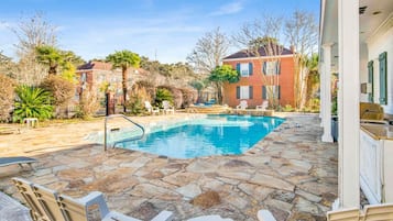 A swimming pool with stone tiles, surrounded by chairs and a building with a red brick facade.
