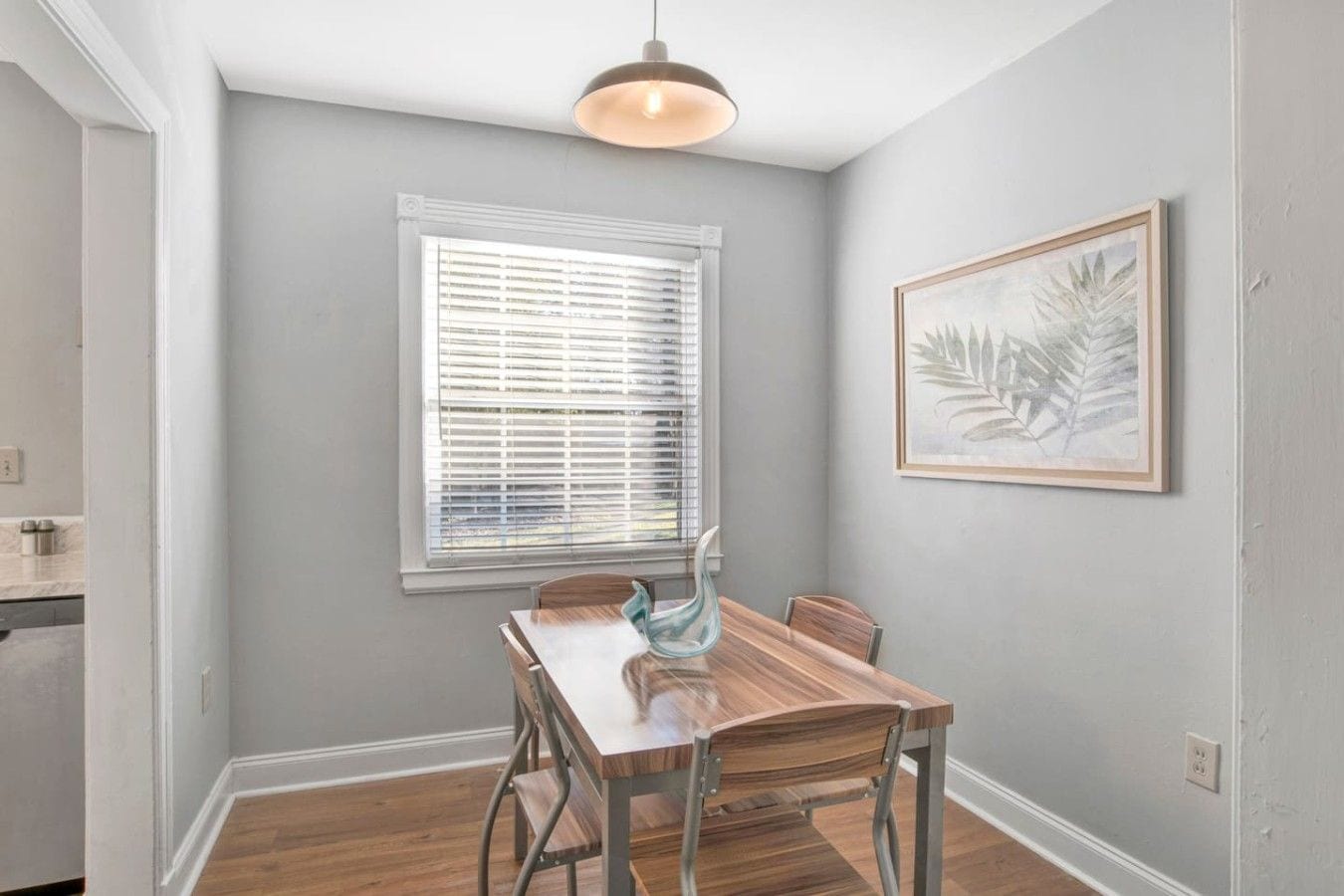 A dining area with a wooden table and chairs, a window with blinds, and a framed picture on the wall.