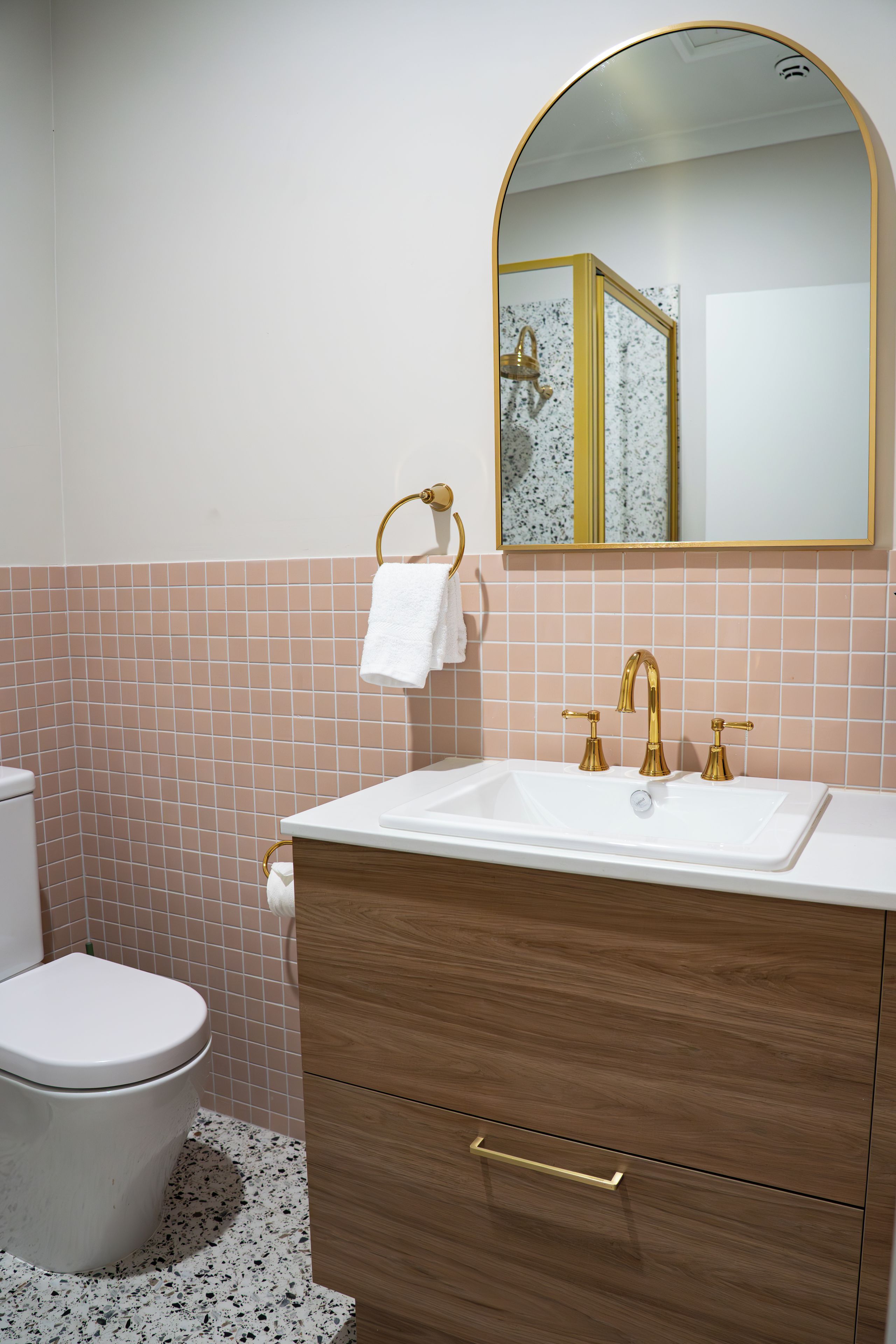 A bathroom with a white sink, gold faucets, a wooden vanity, a white toilet, and a wall-mounted mirror.