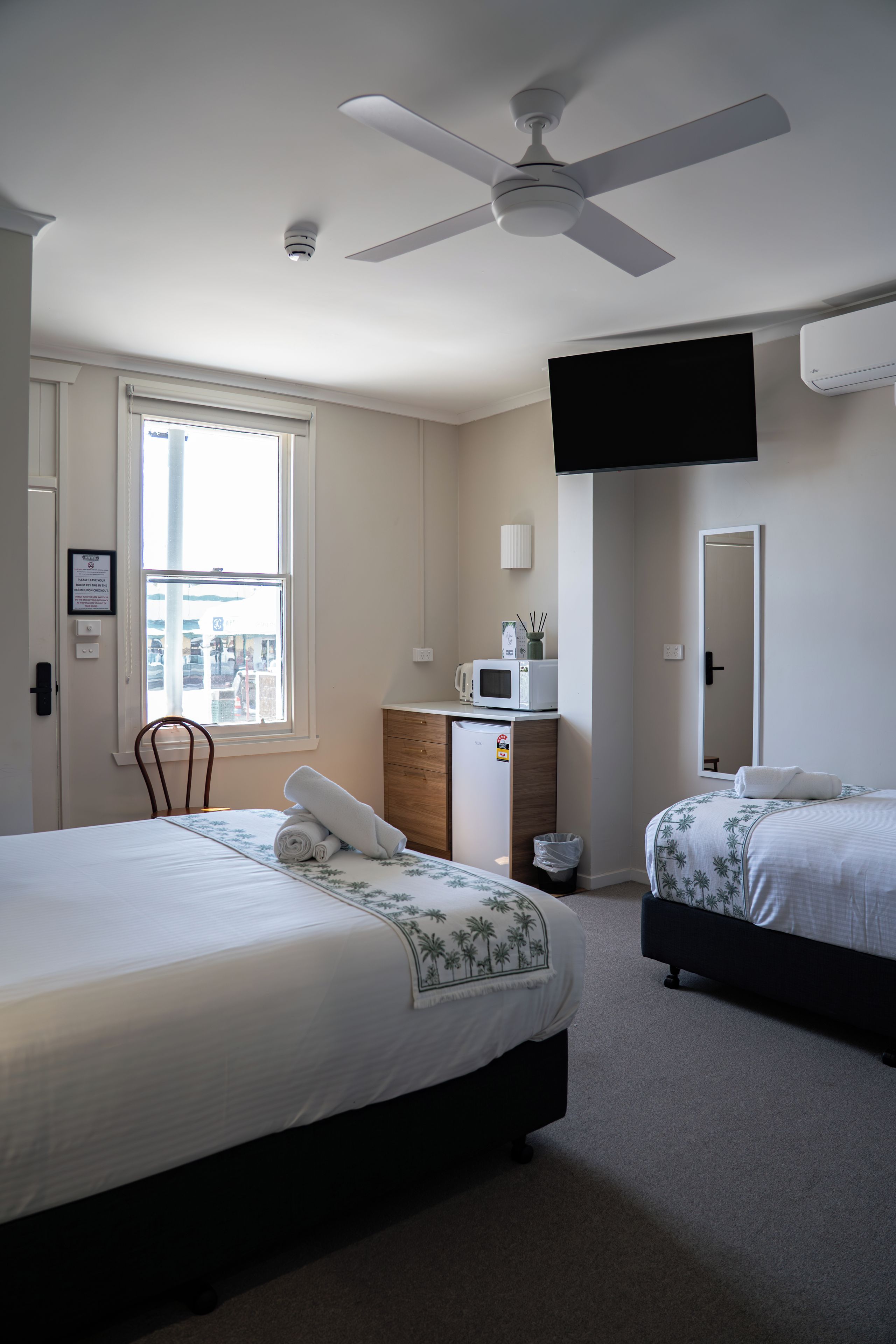 A hotel room with two beds, a ceiling fan, a TV mounted on the wall, a microwave, and a window with a view of the outside.