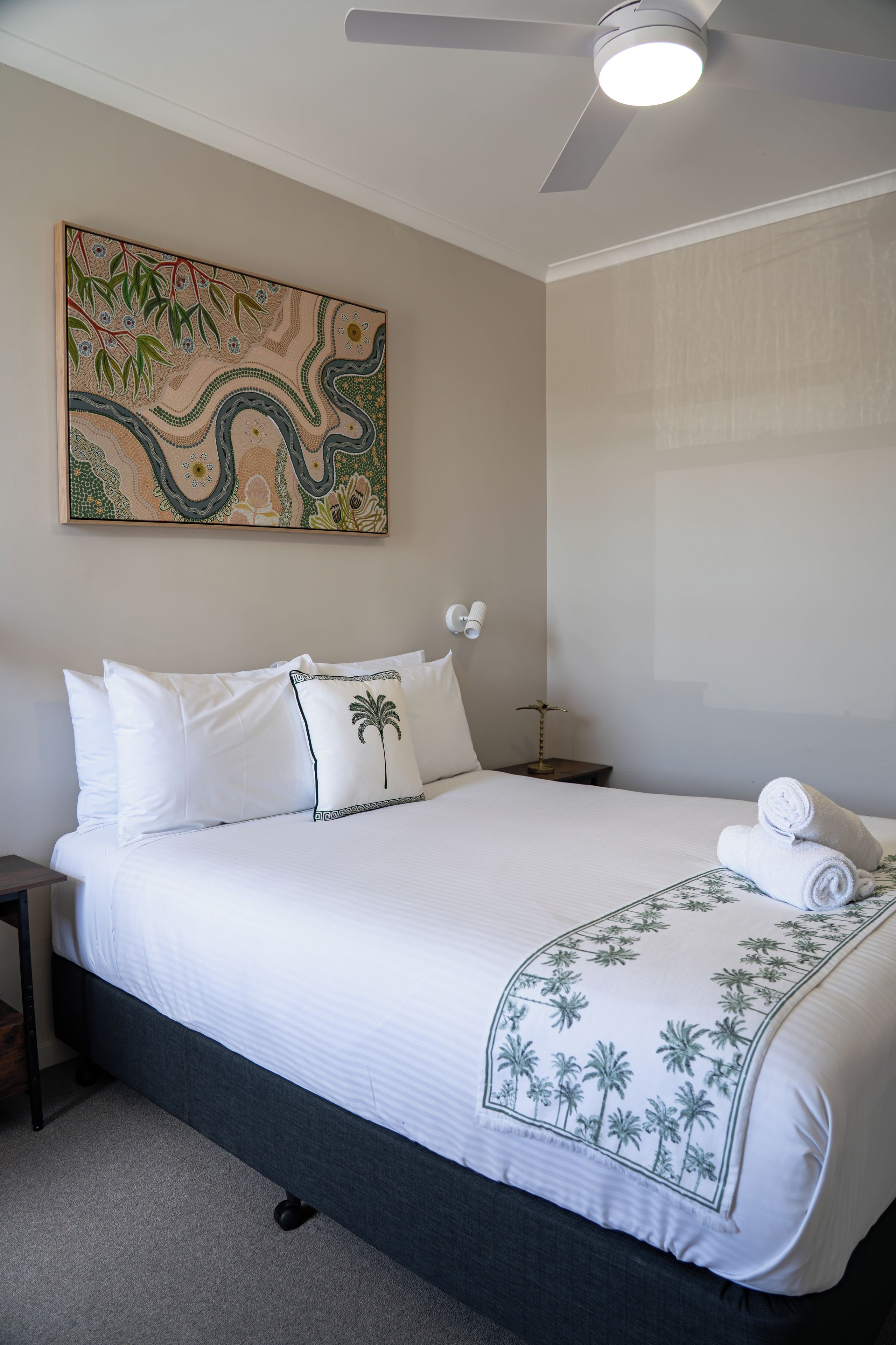 Un lit bien fait, avec du linge de lit blanc orné d’un motif de palmier, un ventilateur de plafond et une œuvre d’art accrochée au mur.