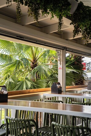 Interior de restaurante con mesas, sillas, plantas colgantes y vistas a palmeras fuera.