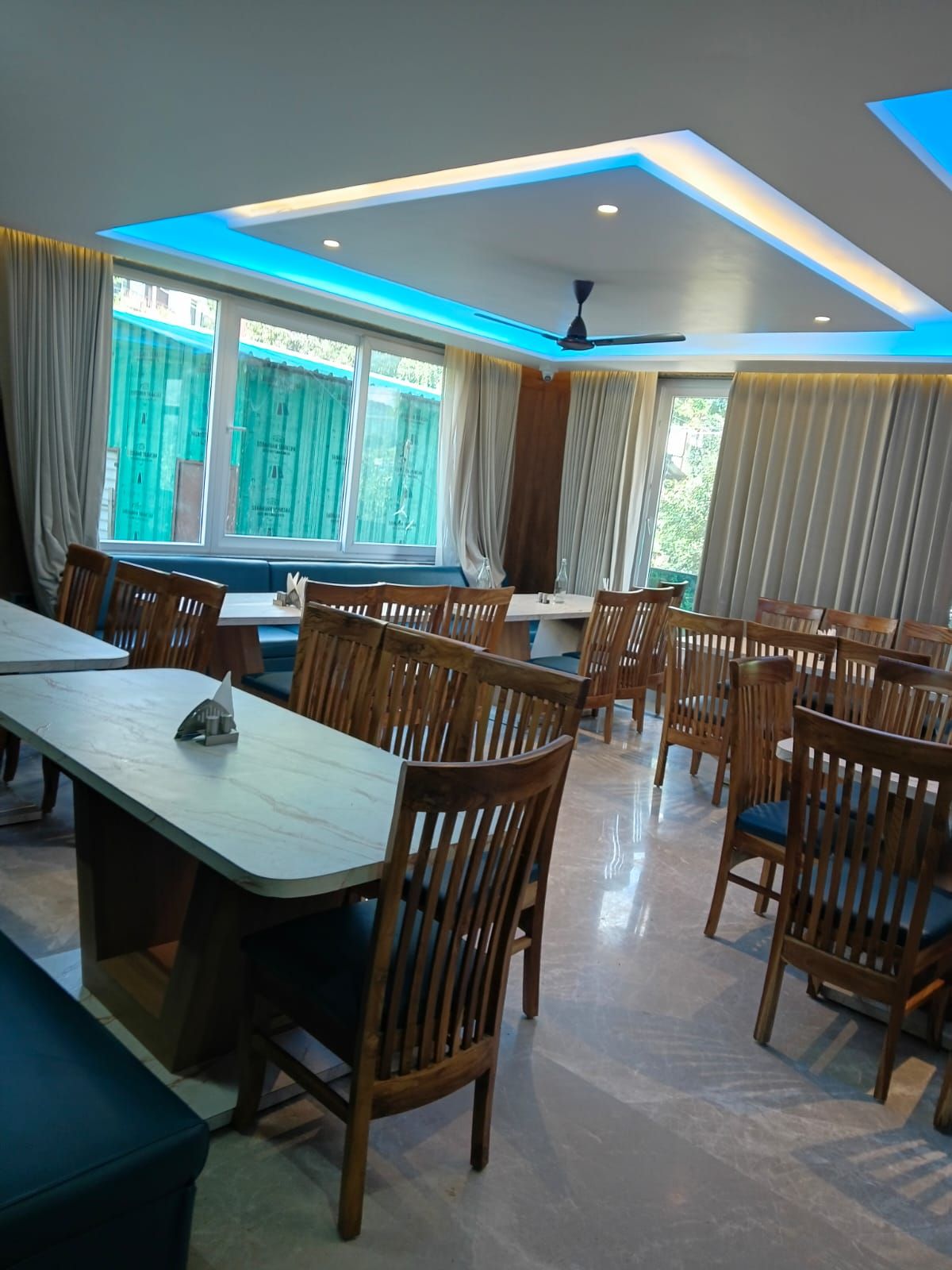 Un comedor con mesas y sillas de madera, ventanas amplias con cortinas e iluminación empotrada en el techo.
