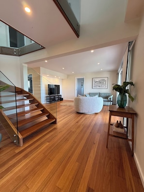 A spacious living area with a wooden staircase, a round seating area, and a dining space. - Love Street by HAUS · 4-Bedroom Kiama Home with Lift, Dog-Friendly (Kiama)