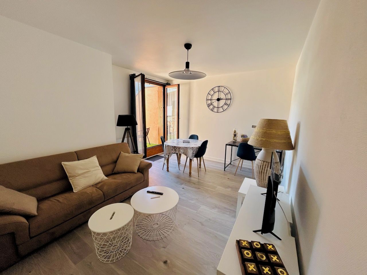 A living room with a brown sofa, two white round tables, a dining table with chairs, a wall clock, and a lamp.