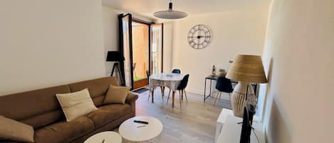 A living room with a brown sofa, two white round tables, a dining table with chairs, a wall clock, and a lamp.