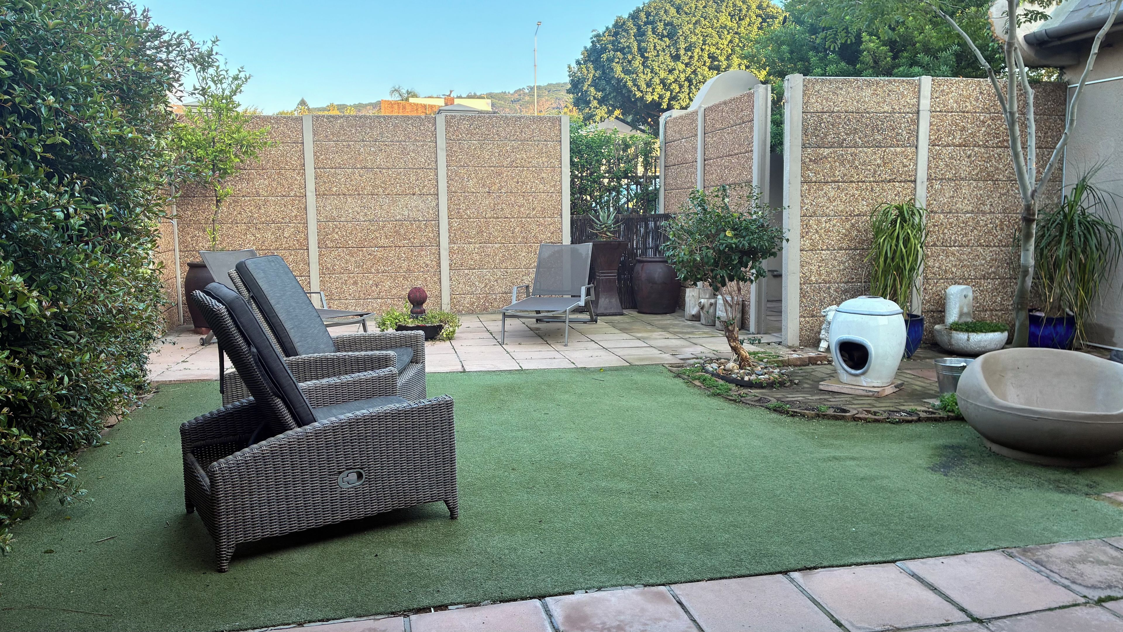 Un patio con césped artificial, una silla de mimbre, un muro de concreto y una maceta grande.