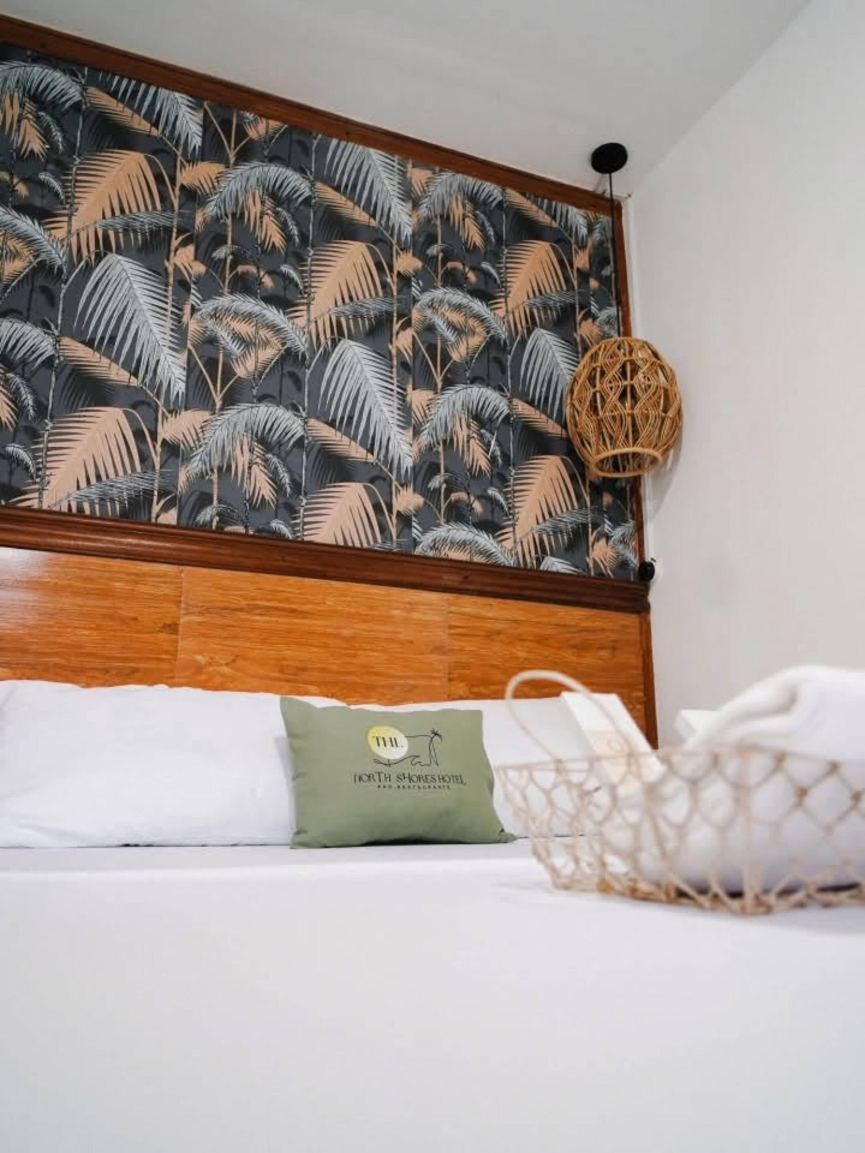 Una cama con cabecera de madera, pared con estampado de hojas tropicales, almohada verde con un logotipo y una cesta tejida.