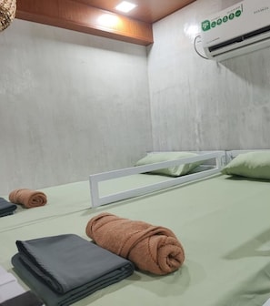 Una cama individual con estructura blanca, colchón verde y almohada. Hay dos toallas enrolladas sobre la cama, una marrón y otra gris.