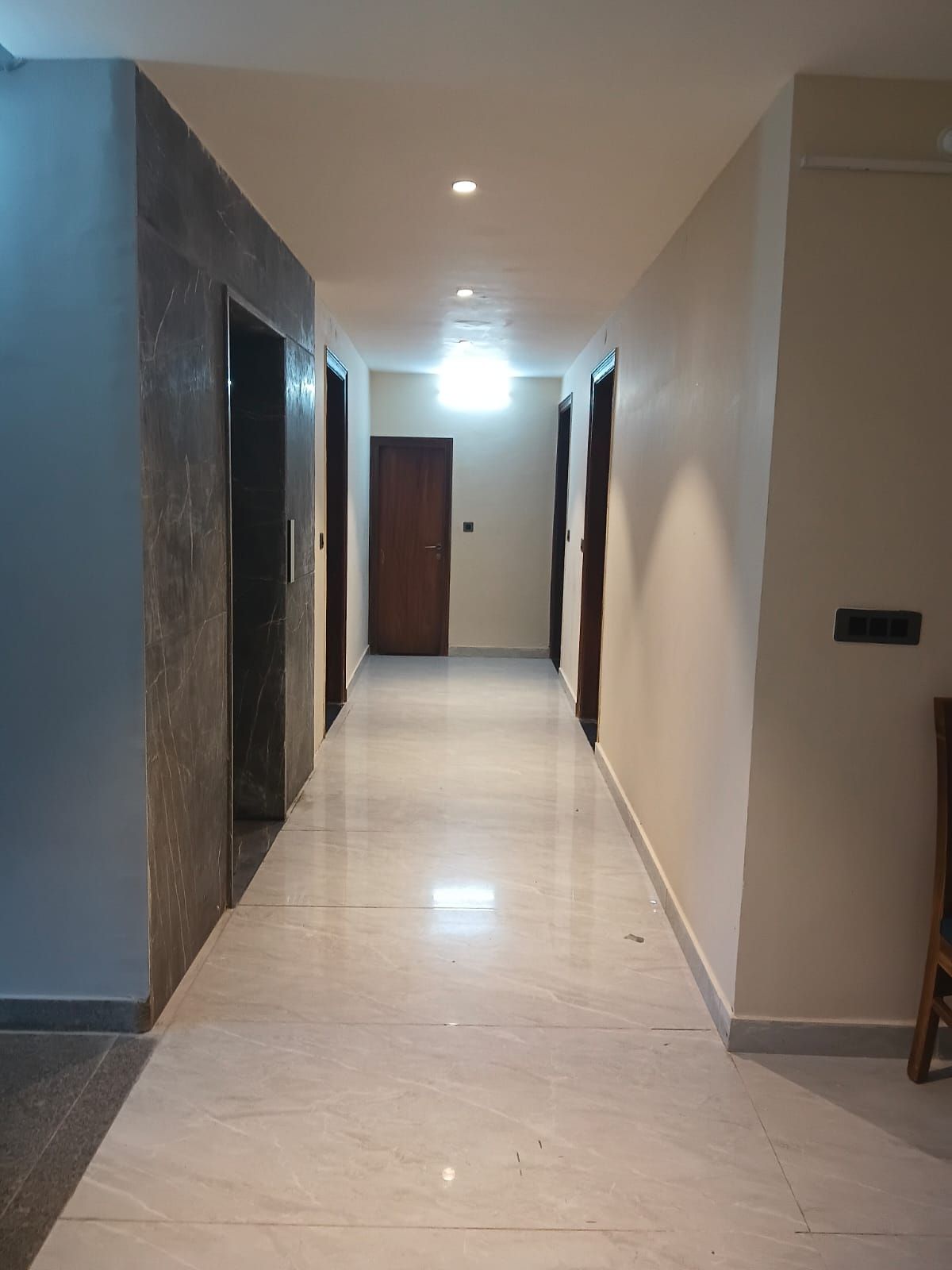 Un pasillo con suelo de baldosas claras, una puerta de madera al final y una lámpara en el techo.
