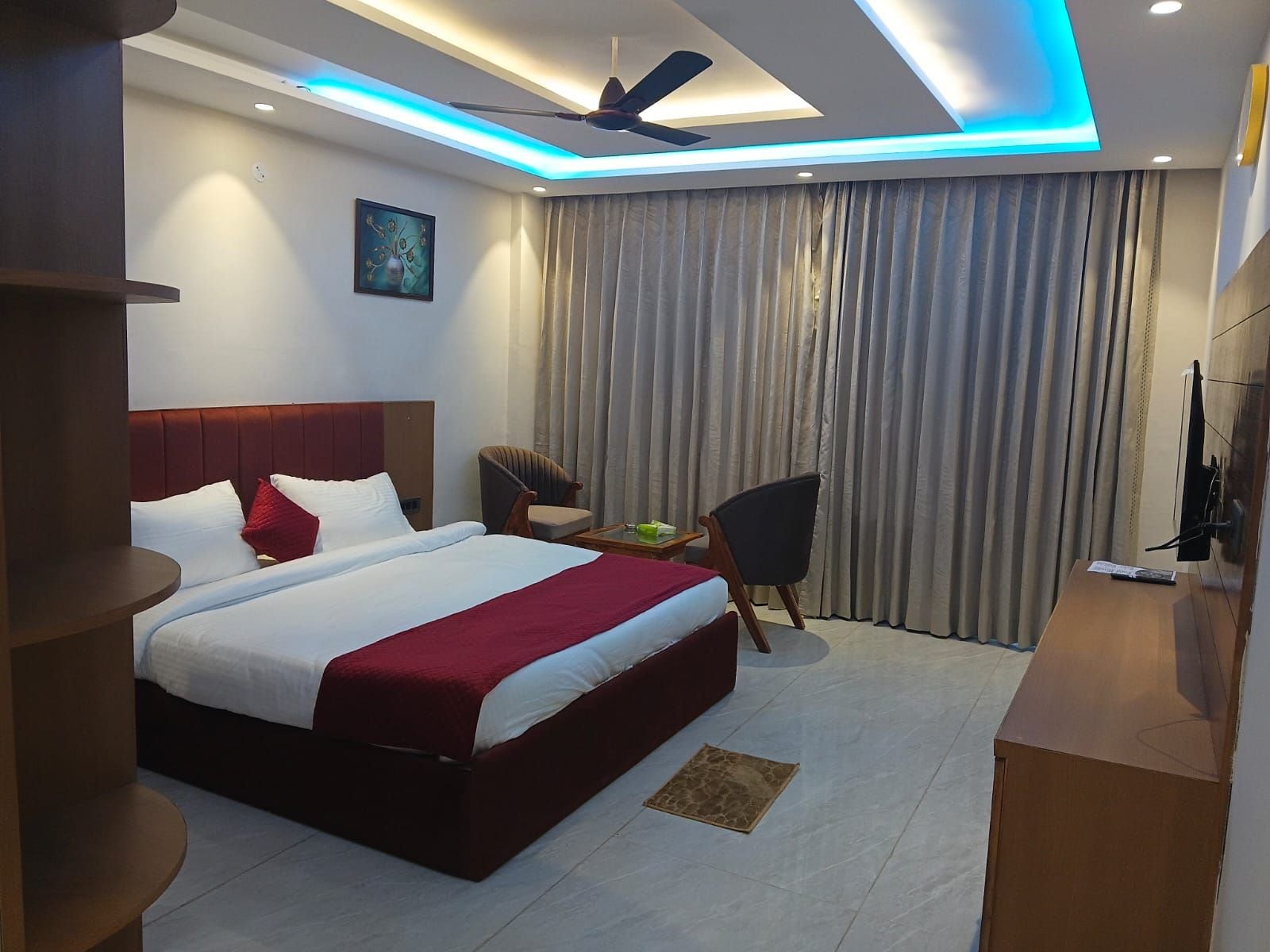 A hotel room with a bed, a desk, a chair, a ceiling fan, and a large window with curtains.