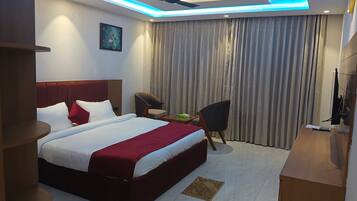 A hotel room with a bed, a desk, a chair, a ceiling fan, and a large window with curtains.