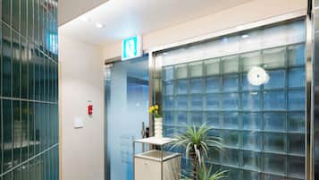 A modern lobby with a reception desk, a potted plant, a glass wall, and a small table with a magazine.