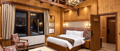 Um quarto amplo com uma cama grande, um espaço de estar com sofá e poltrona, piso de madeira e vigas de madeira aparente no teto.
