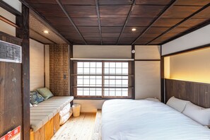 Ein traditioneller japanischer Raum mit Tatami-Boden, einem niedrigen Holzbett, einer eingebauten Sitzbank mit Kissen und einem großen Fenster mit Gittermuster.