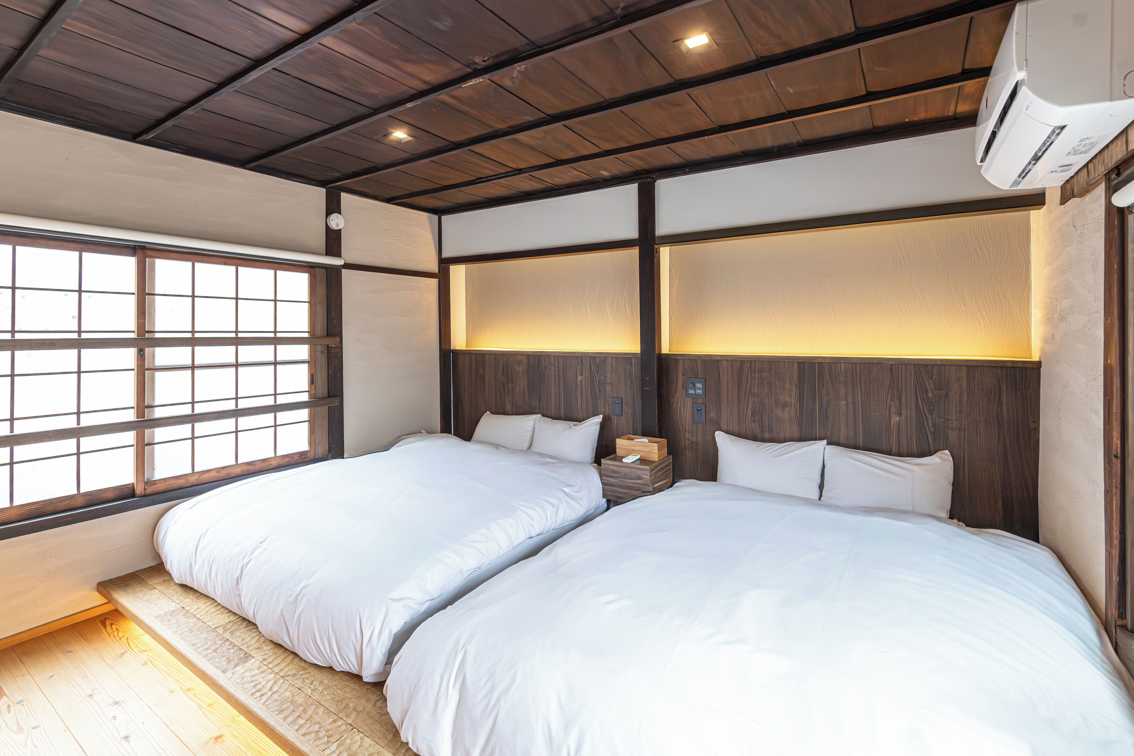 A hotel room with two beds, wooden flooring, a large window with a grid pattern, and a ceiling with wooden beams.
