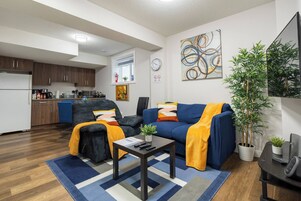 Una sala de estar moderna con sofá, sillón y mesa de centro, una cocina al fondo y un cuadro abstracto grande en la pared.