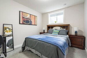 Un dormitorio con cama, mesitas de noche, escritorio, silla, ventana y un cuadro en la pared.