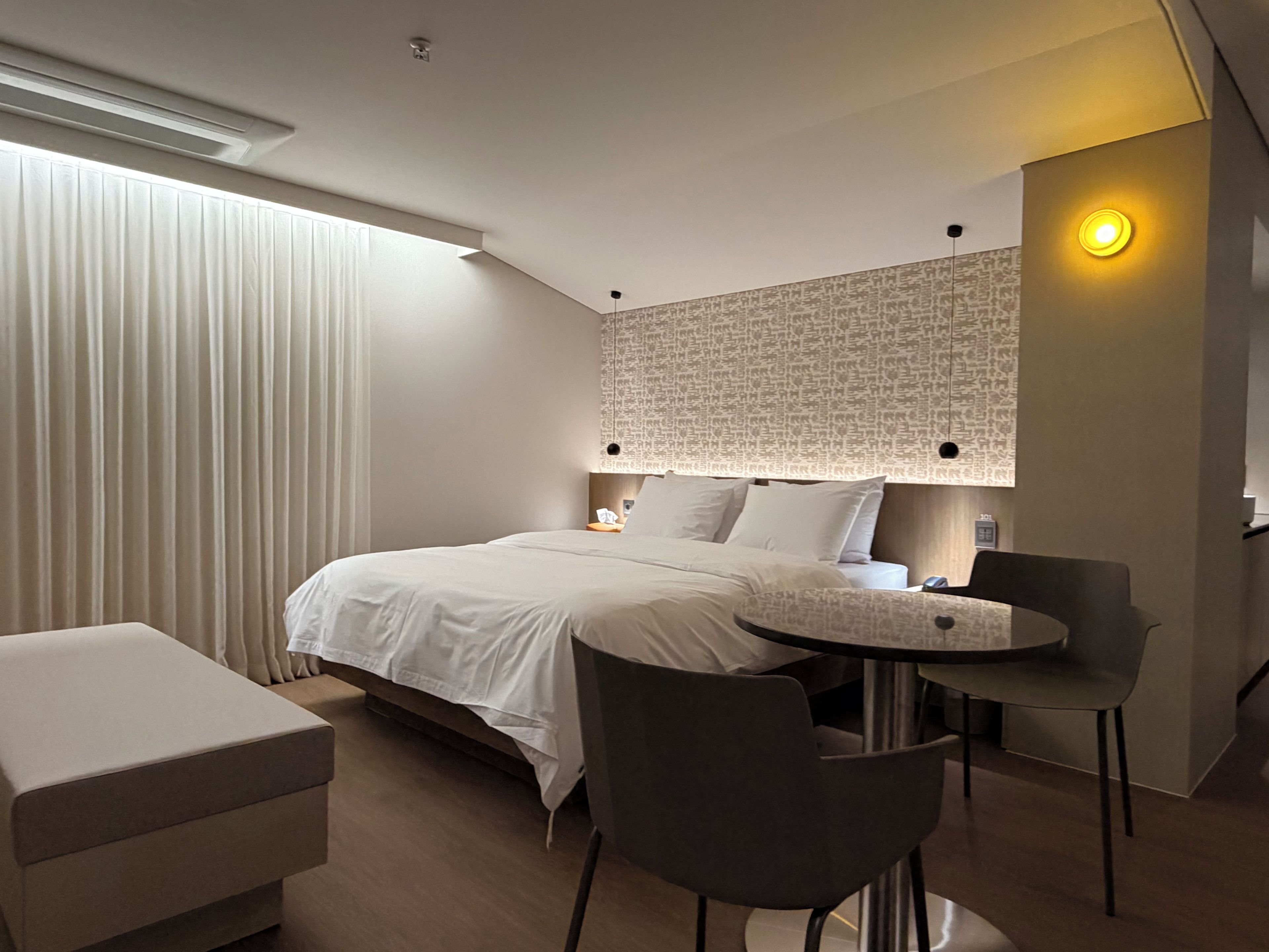 Una habitación de hotel moderna con una cama bien hecha, una mesita redonda con sillas y un ventanal con cortinas.