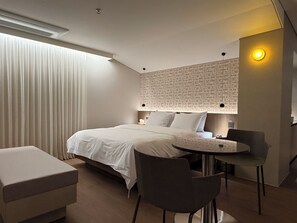 Ein modernes Hotelzimmer mit einem ordentlich bezogenen Bett, einem kleinen runden Tisch mit Stühlen und einem großen Fenster mit Vorhängen.