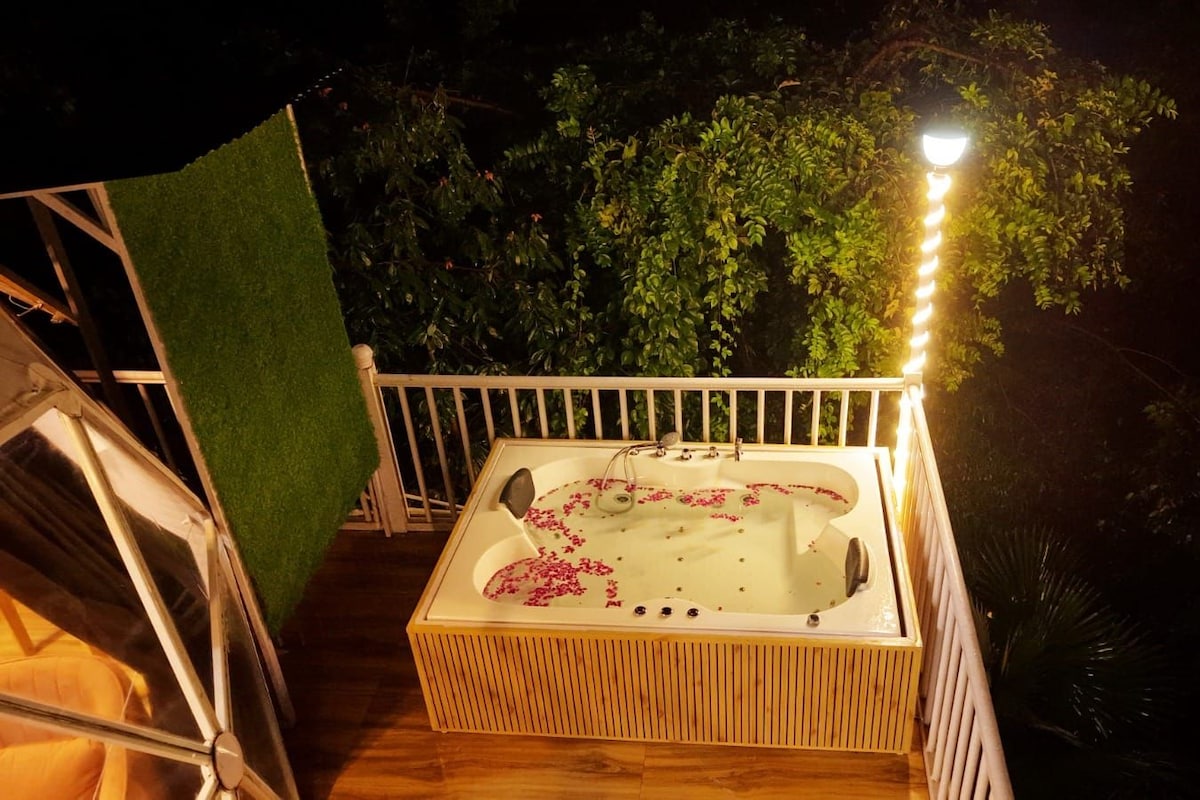 Un jacuzzi en una terraza de madera con barandilla, rodeado de follaje y un cielo nocturno.