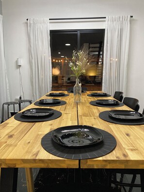 Mesa de comedor dispuesta para ocho personas, con platos negros, un florero transparente con flores y vista a un interior cálidamente iluminado a través de una ventana.