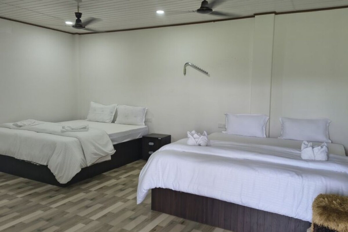 Quarto de hotel com duas camas, um ventilador de teto, uma torneira fixada na parede e piso de madeira.