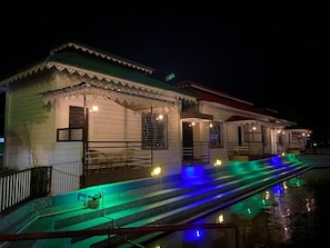 Fila d'edificis amb escales que baixen cap a una superfície aquàtica reflectant, il·luminada per llums interiors.