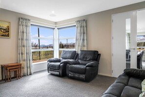 A living room with a sofa, two recliner chairs, a small wooden table, and a view of a landscape through large windows. - The Star Yard - Lake Tekapo (Lake Tekapo)