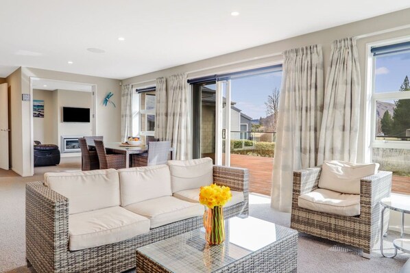 A modern living room with a sofa, a coffee table, and wicker chairs. There is a television and a dining area visible in the background. - The Star Yard - Lake Tekapo (Lake Tekapo)