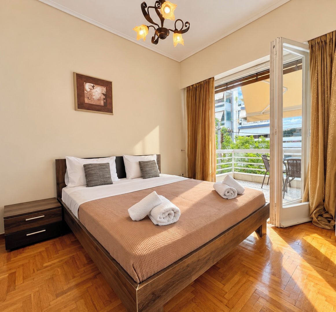 A hotel room with a large bed, a nightstand, a wooden floor, a chandelier, a window with curtains, and a balcony with a view.