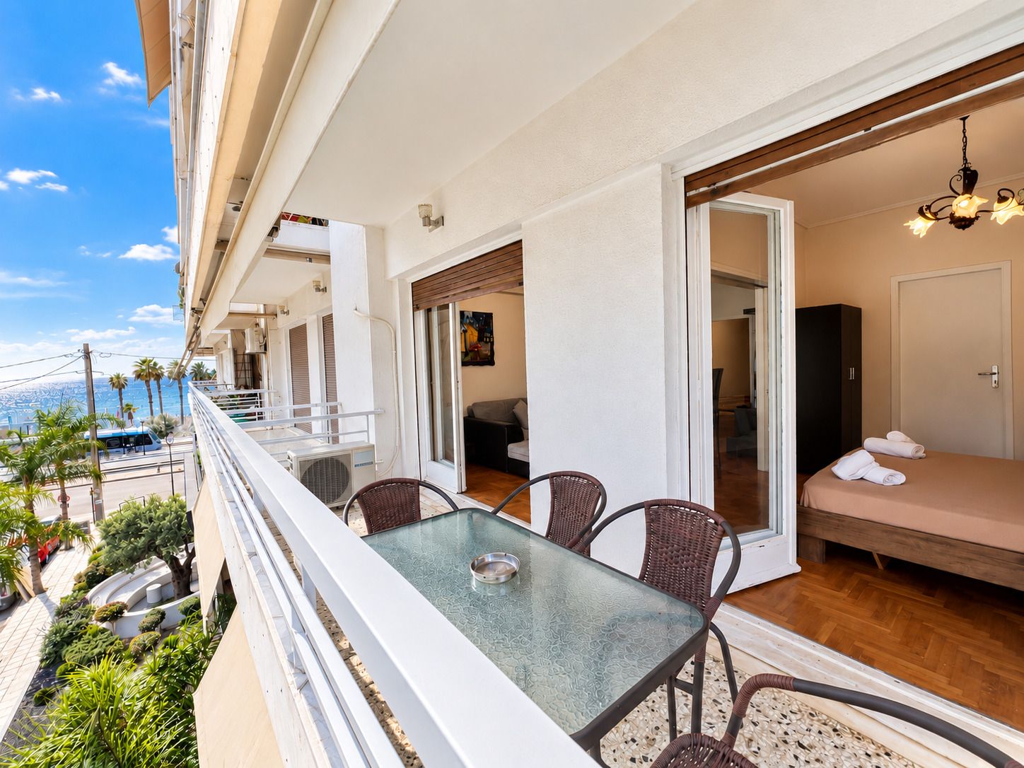 A balcony with a glass table and wicker chairs, overlooking a cityscape with palm trees and a view of the sea.
