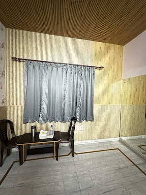 Une pièce avec un plafond à lattes de bois, un rideau gris, une petite table avec une chaise, une chaise noire et un sol carrelé.