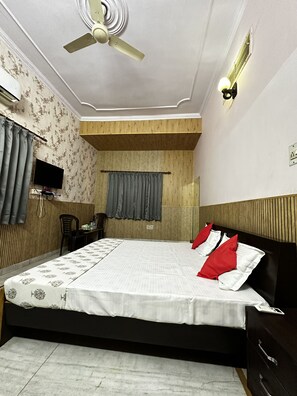 Quarto com cama, ventoinha no teto, luminária na parede, televisão e janela com cortinas.