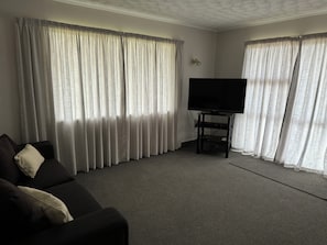 A living room with a black sofa, a flat-screen TV on a stand, and sheer curtains on the windows. - Quaint 3-bedroom house in welcoming Omaha neighborhood (Omaha)