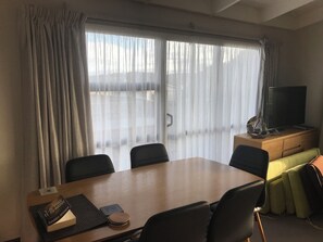 A hotel room with a wooden dining table, black chairs, a television on a wooden cabinet, and a view through a large window with sheer curtains. - Quaint 3-bedroom house in welcoming Omaha neighborhood (Omaha)